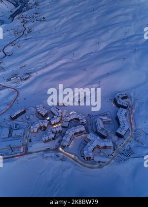 Luftaufnahme der Bergdorf Chalets beleuchtet mit nächtlichen Straßenlaternen entlang Serpentinen kurvige Straße in der Dämmerung bei Sonnenuntergang. Skigebiet mit Schnee Stockfoto