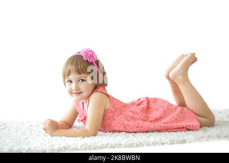 Ein kleines Mädchen in einem rosa Kleid, das auf dem weißen Hintergrund auf dem Teppich liegt Stockfoto