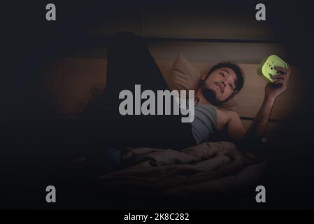 Nachts kann man nicht schlafen, der asiatische junge Mann hält und schaut nachts auf dem Bett auf den Wecker. Stockfoto