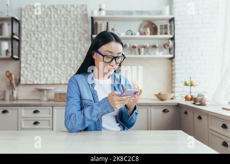 Junge schöne asiatische Frau sitzt in der Küche zu Hause und mit dem Handy. Scrollt durch die Neuigkeiten, sitzt in sozialen Netzwerken, prüft Mails. Stockfoto