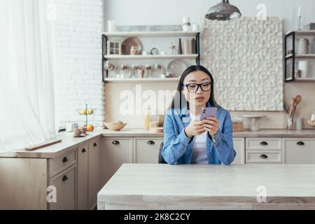 Junge schöne asiatische Frau in Brille und Denim-Kleidung sitzt in der Küche zu Hause und mit einem Handy. Wählt, lächelt. Stockfoto