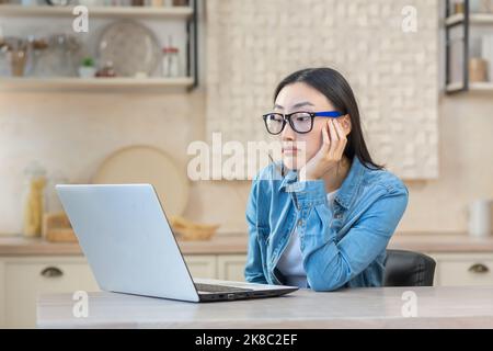 Müde junge asiatische Frau Freiberuflerin sitzt an einem Tisch mit einem Laptop, gelangweilt, nicht in der Lage, Arbeit zu beginnen. Arbeitet während der Quarantäne von zu Hause aus. Stockfoto