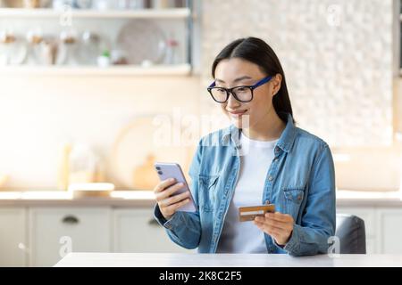 Online-Shopping. Eine lächelnde junge, schöne asiatische Frau macht einen Online-Einkauf und zahlt mit einem bargeldlosen Konto. Er hält ein Telefon und eine Kreditkarte. Sitzen zu Hause in der Küche. Stockfoto