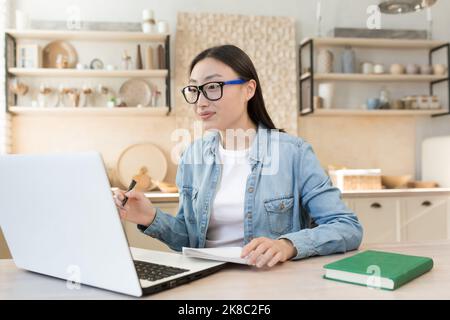 Online lernen. Eine junge Schülerin nimmt per Videoanruf mit einer Lehrerin eine Prüfung aus der Ferne ab. Er sitzt zu Hause in der Küche mit einem Laptop, schaut auf die Kamera, studiert, schreibt in ein Notebook. Stockfoto