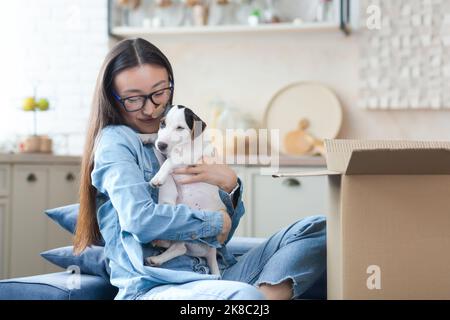 Ein unerwartet angenehmes Geschenk. Eine glückliche und begeisterte junge asiatische Frau erhielt zu ihrem Geburtstag einen kleinen Hund als Geschenk. Er sitzt zu Hause auf dem Sofa in der Nähe eines großen Pappkartons, freut sich und umarmt den Hund. Stockfoto