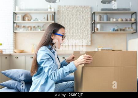 Paketzustellung nach Hause. Glückliche junge schöne asiatische Frau erhielt Paket. Begeistert von ihrer Überraschung öffnet sie einen riesigen Karton, während sie zu Hause auf der Couch sitzt. Stockfoto