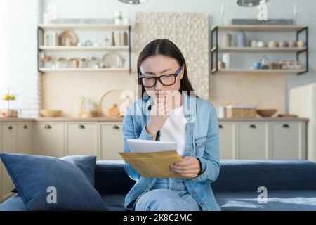 Eine besorgte junge asiatische Frau erhielt einen Brief mit Rechnungen für Kredit, Miete, Hypothek. Zu Hause auf der Couch sitzend, einen Umschlag mit einer Nachricht, Quittungen und Schulden haltend. Stockfoto