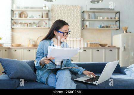 Online zu Hause arbeiten. Junge asiatische Buchhalterin, die zu Hause arbeitet. Sitzen auf der Couch mit einem Laptop und Dokumenten. Stockfoto