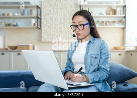 Online arbeiten. Eine junge Asiatin ist Callcenter-Betreiber. Sitzen auf dem Sofa zu Hause in Kopfhörer mit Mikrofon und Laptop, Anrufe entgegennehmen, beraten, schreiben eine Anfrage. Stockfoto