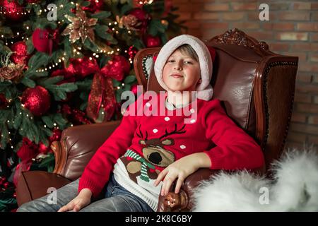 Ein aufgebrachter Junge in einem alten Stuhl neben einem Weihnachtsbaum mit roten Bällen, ein Junge mit Weihnachtsmann-Hut, der an Weihnachten in einem Hirsch-Pullover auf Geschenke wartet Stockfoto