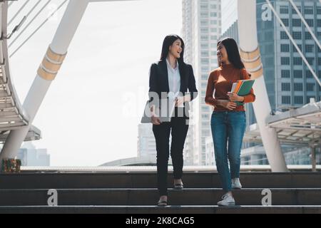 Glückliche zwei Teenager High School Schülerinnen gehen und sprechen zusammen, tragen Laptop und Bücher in der Hand, Outdoor-Bildungskonzept. Stockfoto