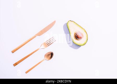 Avocado und Besteck auf weißem Hintergrund. Gesundes veganes Rohkost-Konzept. Scharfe Schatten. Draufsicht, flach liegend. Stockfoto