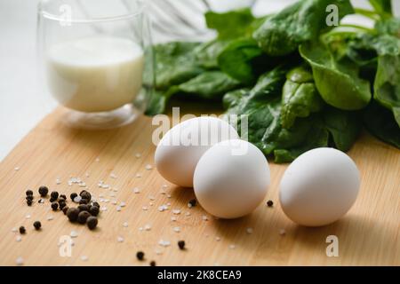 Drei weiße Hühnereier auf einem Schneidebrett mit einem Haufen frischem Spinat. Ein Glas Milch im Hintergrund. Stockfoto