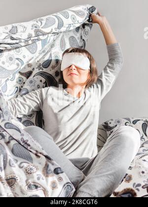 Junge Frau in grauem Pyjama und Schlafmaske im Gesicht. Sie ist gerade aufgewacht und sitzt im Bett. Am frühen Morgen in gemütlichem Zuhause. Stockfoto