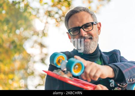 Ein lächelnder, eleganter, bärtiger, grauhaariger, reifer Mann mit Brille auf, hält ein rotes Penny-Brett und berührt seine Räder. Hochwertige Fotos Stockfoto