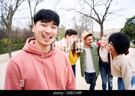 Junger lächelnder asiatischer Kerl, der mit einer Gruppe von Freunden die Kamera im Freien anschaut. Glückliche Menschen gemeinsam Spaß haben Konzentrieren Sie sich auf einen jungen Chinesen Stockfoto