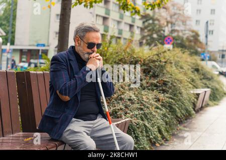 Blinder bärtiger grauhaariger reifer Mann mit dunkler Brille und einer Jacke, die einen Spazierstock hält und auf einer Bank im Park sitzt. Hochwertige Fotos Stockfoto