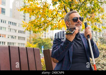 Eleganter, blinder, grauhaariger, reifer Mann mit Bart und dunkler Sonnenbrille, der einen Spazierstock hält, die Herbstsaison im Park genießt und über ein Freisprechtelefon mit jemandem spricht. Hochwertige Fotos Stockfoto