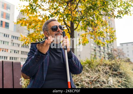 Eleganter, blinder, grauhaariger, reifer Mann mit Bart und dunkler Sonnenbrille, der einen Spazierstock hält, die Herbstsaison im Park genießt und mit jemandem über eine Freisprecheinrichtung spricht. Hochwertige Fotos Stockfoto