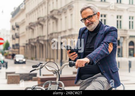 Moderner bärtiger grauhaariger reifer Mann, der sein Smartphone benutzt und mit dem Fahrrad in den Straßen der Stadt mit schönen Gebäuden im Hintergrund fährt. Hochwertige Fotos Stockfoto