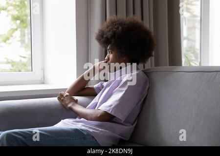 Trauriger afrikanischer Teenager sitzt auf dem Sofa und schaut durch das Fenster Stockfoto