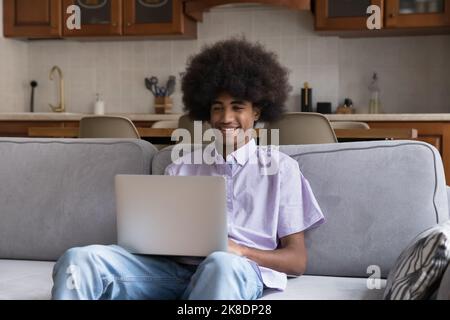 Ein gutaussehender, lächelnder Teenager sitzt mit einem Laptop auf dem Sofa Stockfoto