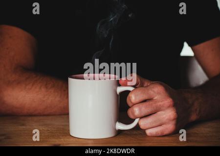 Mann hält dampfenden Becher auf einem Holztisch Stockfoto