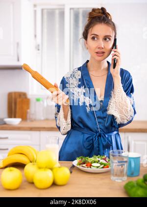 Unzufriedene Hausfrau mit einem Nudelholz in den Händen, die in der Küche auf dem Mobiltelefon sprach Stockfoto