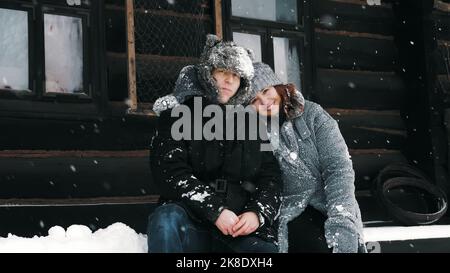 Winterporträt. Fröhliches, lustiges Paar verliebt, Mann und Frau, in warme Winterkleidung gekleidet, voll mit Schneeflocken bedeckt, sitzen auf der Bank bei einem alten Holzhaus und umarmen. Glückliche Zeit am verschneiten Wintertag. Hochwertige Fotos Stockfoto