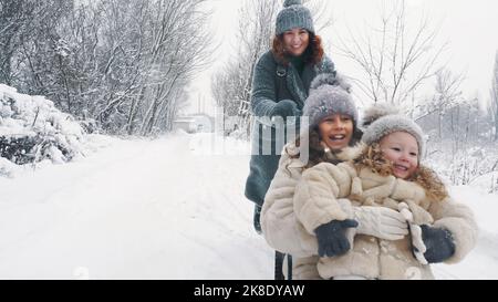 Familienrodeln im Winter. Outdoor Winteraktivitäten. Glückliche, lachende Familie, Frau mit 2 Töchtern genießen Schlittenfahrten auf verschneiten Straßen, im Wald, bei Schneefall. Familie hat Spaß, Zeit zusammen am verschneiten Wintertag zu verbringen. Zeitlupe. Hochwertige Fotos Stockfoto
