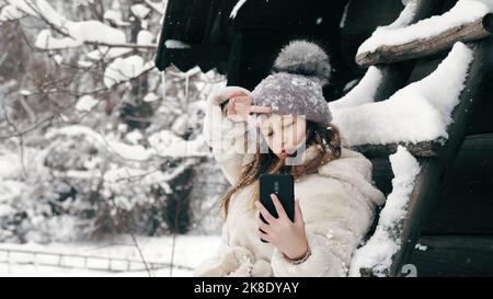Verschneite Winter-Selfie. Cutie-Mädchen, gekleidet in warme Winterkleidung, voll mit Schneeflocken bedeckt, sitzt auf der Bank und nimmt Selfie, mit dem Smartphone, im Freien, während Schneefall, in der Nähe von alten Holzhaus. Verschneiten Wintertag. Hochwertige Fotos