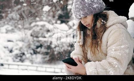 Winterporträt. Cutie kleines Mädchen, in warmen Winterkleidung gekleidet, voll mit Schneeflocken bedeckt, sitzt auf der Bank und mit dem Smartphone, im Freien, während Schneefall, in der Nähe von alten Holzhaus. Verschneiten Wintertag. Hochwertige Fotos