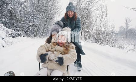 Familienrodeln im Winter. Outdoor Winteraktivitäten. Glückliche, lachende Familie, Frau mit 2 Töchtern genießen Schlittenfahrten auf verschneiten Straßen, im Wald, bei Schneefall. Familie hat Spaß, Zeit zusammen am verschneiten Wintertag zu verbringen. Zeitlupe. Hochwertige Fotos Stockfoto