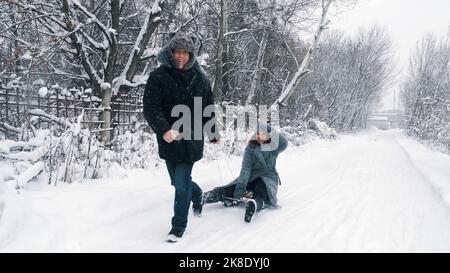 Familienrodeln im Winter. Outdoor Winteraktivitäten. Glückliches, lachendes, verspieltes Ehepaar genießt das Rodeln auf verschneiten Straßen, im Wald, bei Schneefall. Sie haben Spaß, verbringen Zeit zusammen am verschneiten Wintertag. Zeitlupe. Hochwertige Fotos Stockfoto