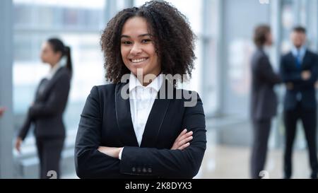 Selbstbewusste Geschäftsfrau, die die Kamera anschaut, weibliche Teamleiterin, die für Fotos posiert, im modernen Büro mit gekreuzten Armen und einem vielfältigen Team auf dem Rücken steht Stockfoto