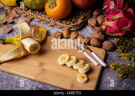 Banane kanarischen Ursprungs auf einem Holzbrett in Scheiben geschnitten, begleitet von tropischen Früchten wie Pitahaya, Kakimmon und Pudding Apple. Stockfoto