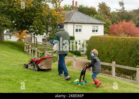 Vater und Sohn mähen das Gras, der niedliche Junge schiebt einen Spielzeug-Rasenmäher mit Ohrenschützern und imitiert und folgt seinem Vater, Großbritannien Stockfoto