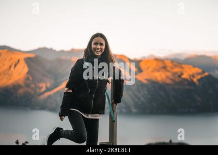 kaukasische niedliche Brünette Mädchen in schwarzer Jacke schwarze Hosen und Sneakers halten auf Holzstab mit Knie angehoben und auf einem Bein glücklich und stehen Stockfoto