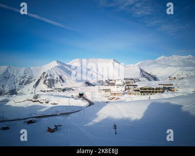 Luftaufnahme der verschneiten Bergkette bei Wintersonnenaufgang im Skigebiet. Drohne über Bergtal und Dorf mit kurviger Straße bei Sonnenuntergang. Gipfel des Kaukasus Stockfoto