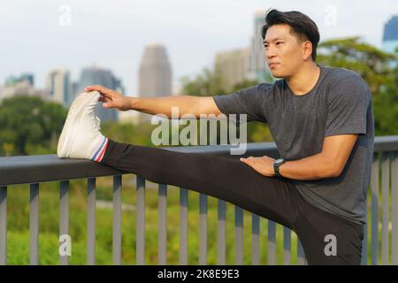 Stadtsport - Asiatischer junger Mann macht sich warm, bevor er an einem schönen Sommertag in der Stadt läuft Stockfoto