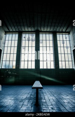 Eine einzige Bank in einem alten Waschraum. Das Licht scheint durch die Fenster. Der ganze Raum ist mit Fliesen gefliest und unter der Decke hängt noch alter Draht Stockfoto