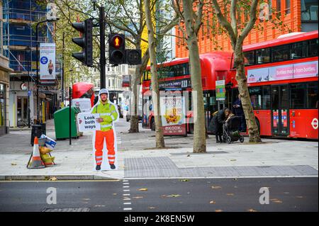 LONDON - 3. November 2020: Schauen Sie in beide Richtungen auf ein ausgeschnittenes Kartonschild des Arbeiters am Fußgängerübergang mit einer roten Mann-Stoppleuchte. Roter London Doppeldecker Stockfoto