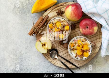 Gesundes Frühstücksgranola im Herbst oder Winter. Hausgemachtes Dessert mit Joghurt, Müsli, Karamelläpfeln und Kürbis, Zimt, Nüssen auf einem Steintisch. Ansicht f Stockfoto