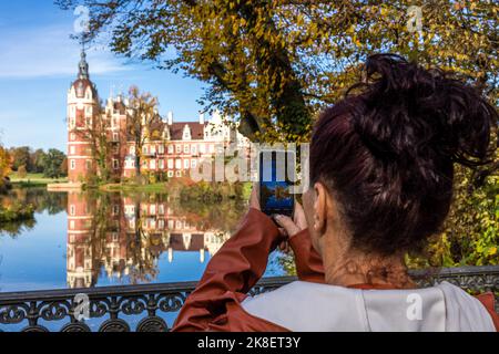 Bad Muskau, Deutschland. 23. Oktober 2022. Eine junge Frau fotografiert das Neue Schloss, das sich in einem Teich im Prinz-Pückler-Park spiegelt, mit ihrem Smartphone im Sonnenschein. Ein beliebtes Ausflugsziel ist der rund 800 Hektar große Landschaftspark im englischen Stil in der Oberlausitz. Quelle: Frank Hammerschmidt/dpa/Alamy Live News Stockfoto