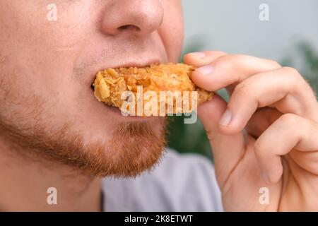 Ein Mann beißt und kaut einen gebratenen Hühnerflügel in Brotkrumen. Ein Mann isst Fast Food. Ungesunde Ernährung, schlechte Gewohnheiten. Stockfoto