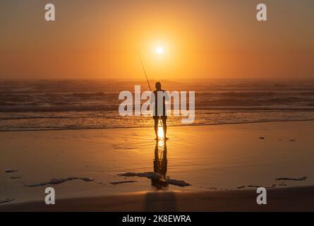 Ein alleinstehender Fischer steht an der Küste, reflektiert in nassem Meeressand und beleuchtet mit dramatischem roten Sonnenuntergangslicht Stockfoto