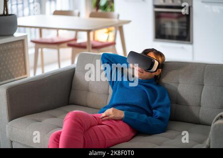 Frau, die auf einer bequemen Couch sitzt, spielt gerne in einer VR-Brille in der Wohnung. Cyberspace Stockfoto