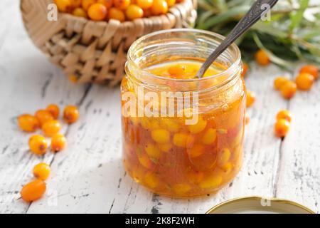 Krug mit gesunder Sanddornmarmelade auf dem weißen Tisch. Korb mit Sanddornbeeren auf dem Hintergrund. Stockfoto