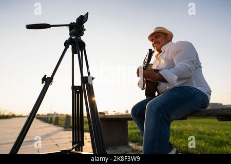 Lächelnder Straßenmusiker, der Gitarre spielt und über ein Smartphone auf einem Stativ aufnimmt Stockfoto