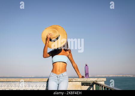 Junge Frau bedeckt das Gesicht mit einem Hut, der an einem sonnigen Tag an der Wand steht Stockfoto